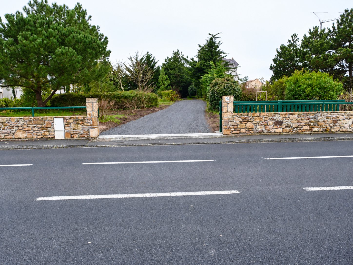 Une allée de gravier fraîchement réalisée mène à une propriété, entre deux murets en pierre surmontés de grilles vertes, face à une route.