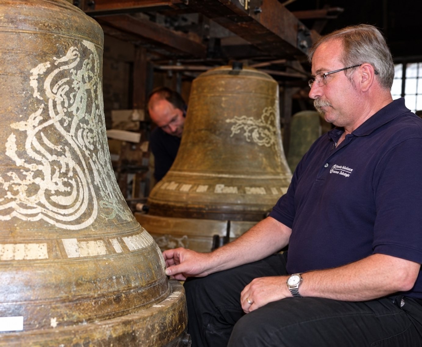 Un homme en polo sombre pose la main sur une grande cloche en bronze décorée de motifs, d'autres cloches et une personne visibles derrière lui.