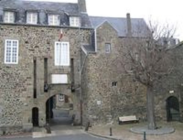Un ancien bâtiment en pierre grise avec une arche et des fenêtres blanches, près d'un arbre sans feuilles et d'un banc sur une petite place.