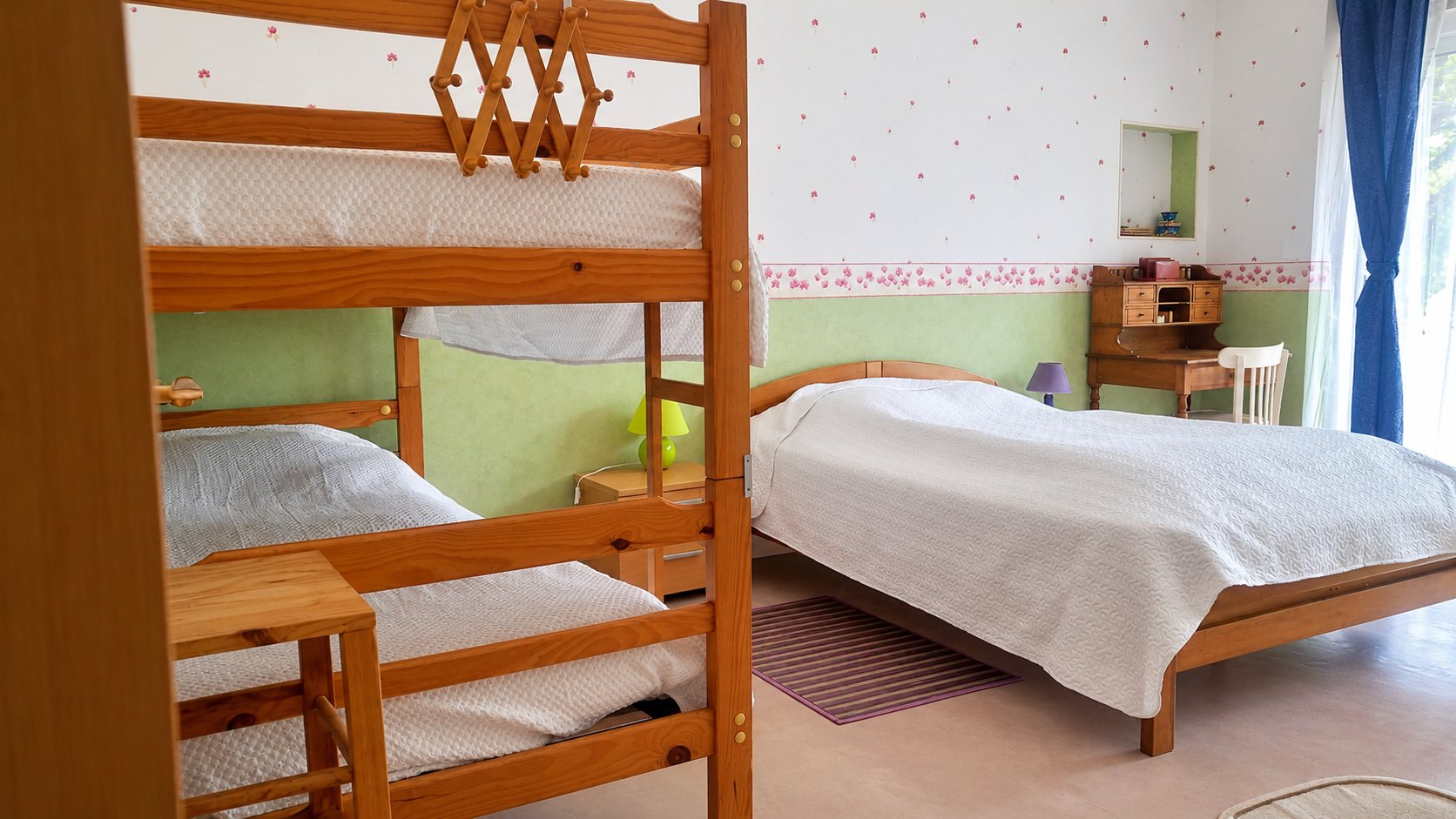 Chambre aux murs vert pâle et blancs avec des lits superposés en bois, un grand lit double, un petit bureau ancien et un rideau bleu.
