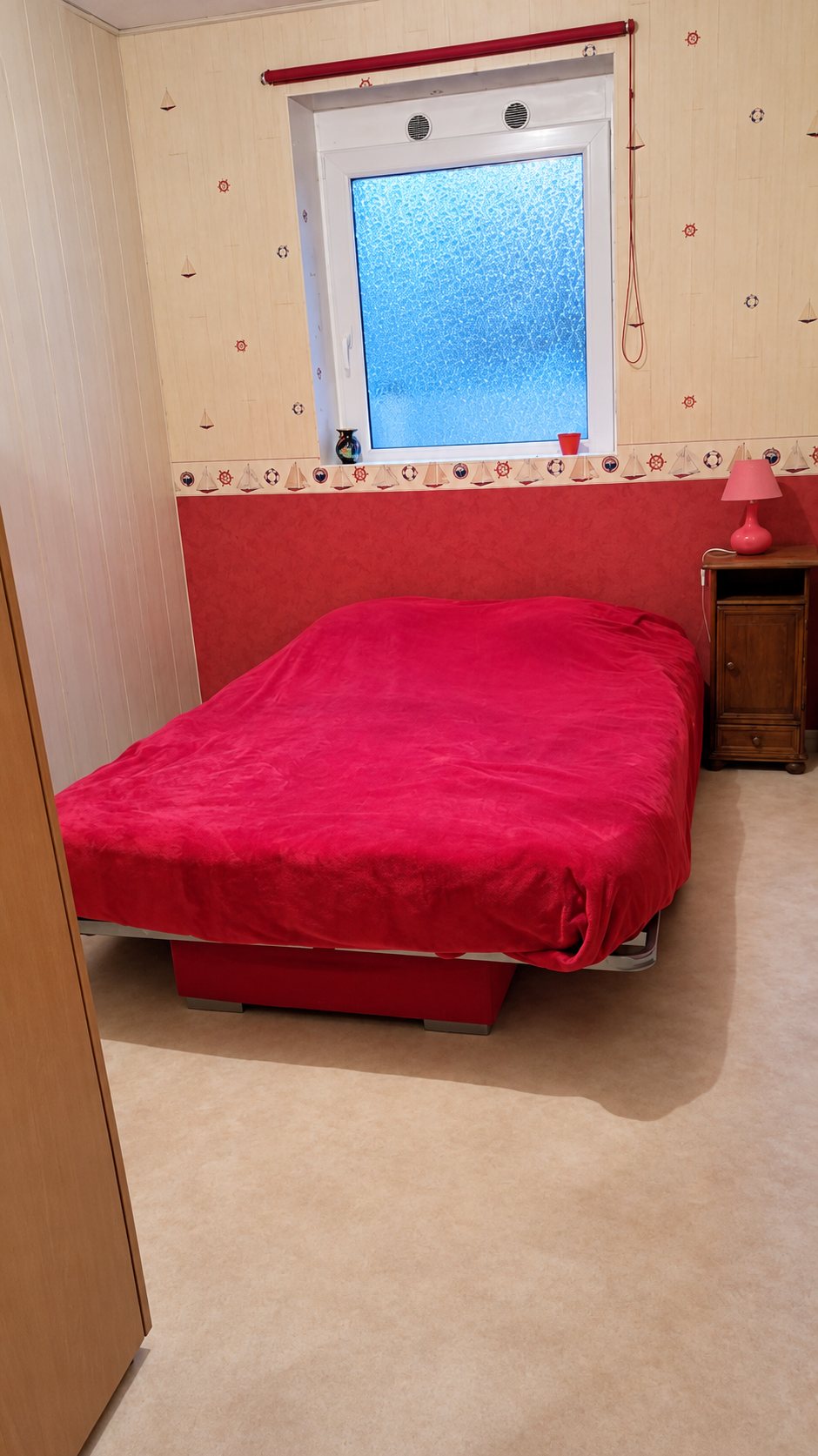 Une chambre aux murs rouges et crème avec un lit double couvert d'un plaid rouge, une fenêtre en verre dépoli et une table de chevet en bois avec une lampe rose.