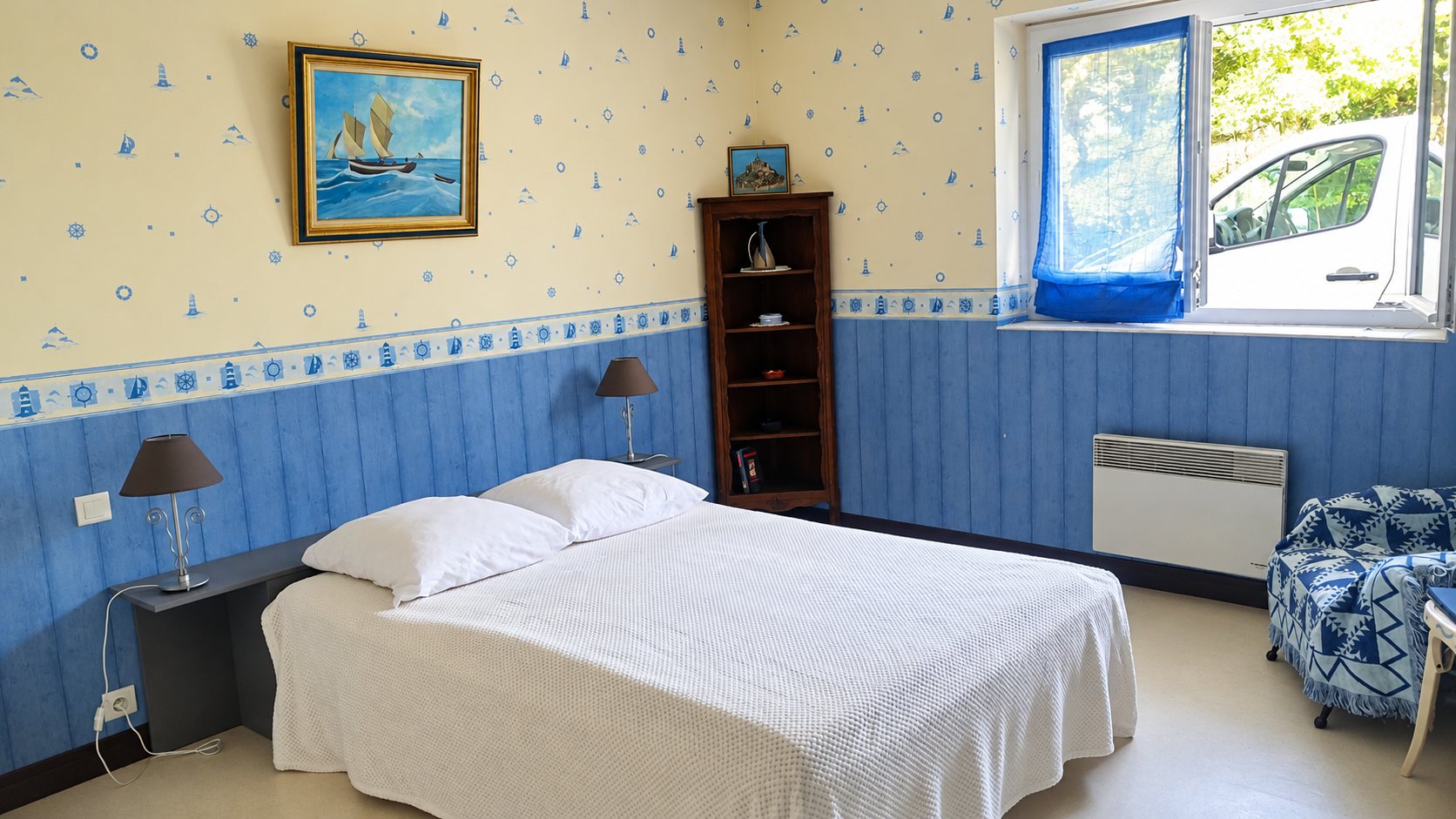 Chambre avec un grand lit double au couvre-lit blanc, des lambris bleus, un papier peint à motifs marins, un tableau de voilier et une fenêtre ouverte.