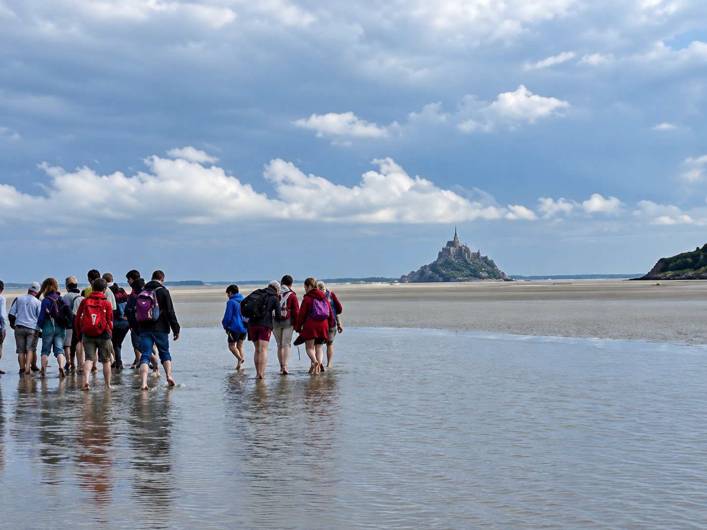 Un groupe de randonneurs avec sacs à dos marche pieds nus sur le sable mouillé de la baie, avec le Mont-Saint-Michel au loin sous un ciel nuageux.