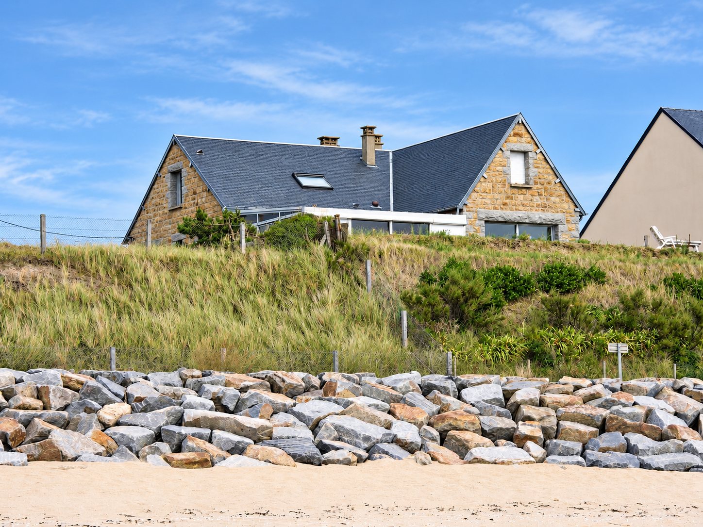 Une maison en pierre au toit d'ardoise domine une dune herbeuse, protégée par un mur d'enrochements de gros blocs de granit au-dessus d'une plage de sable.