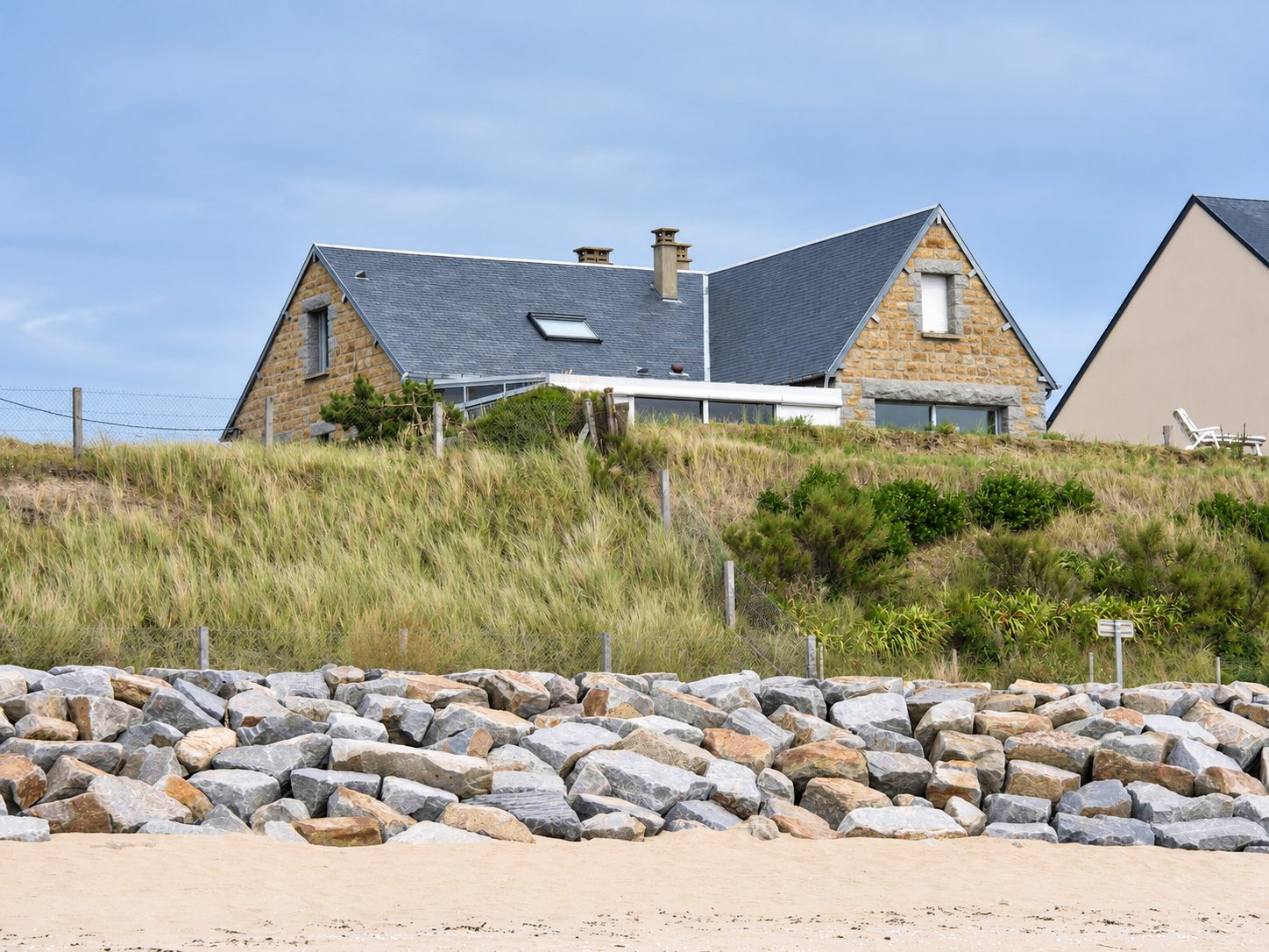 Une maison en pierre au toit d'ardoise domine une dune herbeuse, protégée par un mur d'enrochements de gros blocs de granit au-dessus d'une plage de sable.