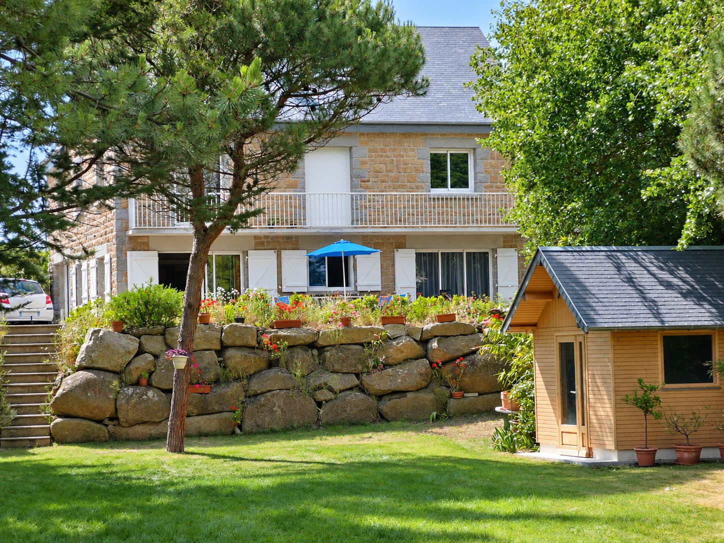Une maison en pierre avec balcon et terrasse fleurie surplombe un muret de gros rochers, un pin, une pelouse et un petit abri de jardin en bois.