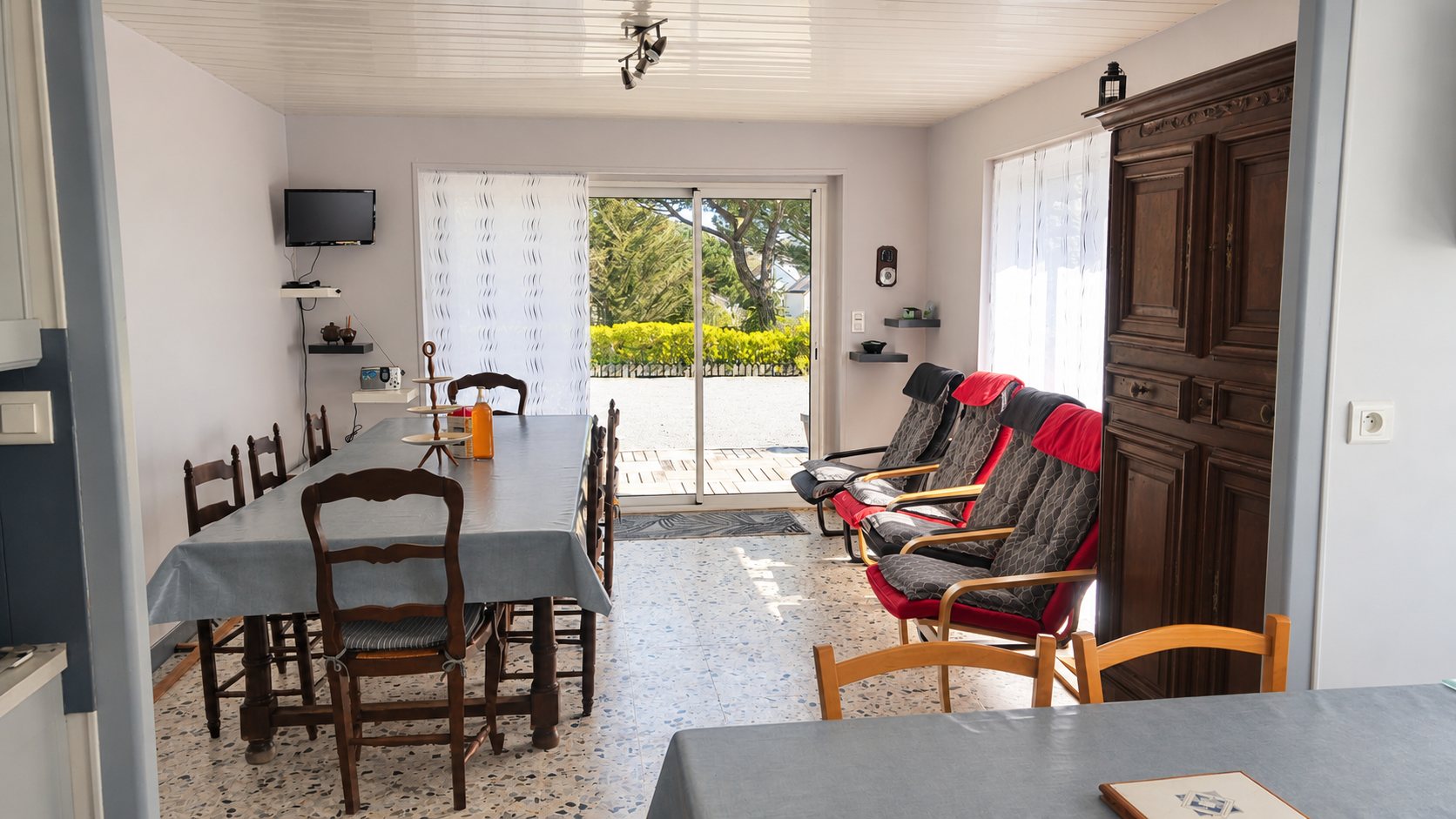 Pièce à vivre lumineuse avec une longue table recouverte d'une nappe, des chaises en bois, des fauteuils rouges et une grande armoire ancienne, ouverte sur un jardin par une baie vitrée.