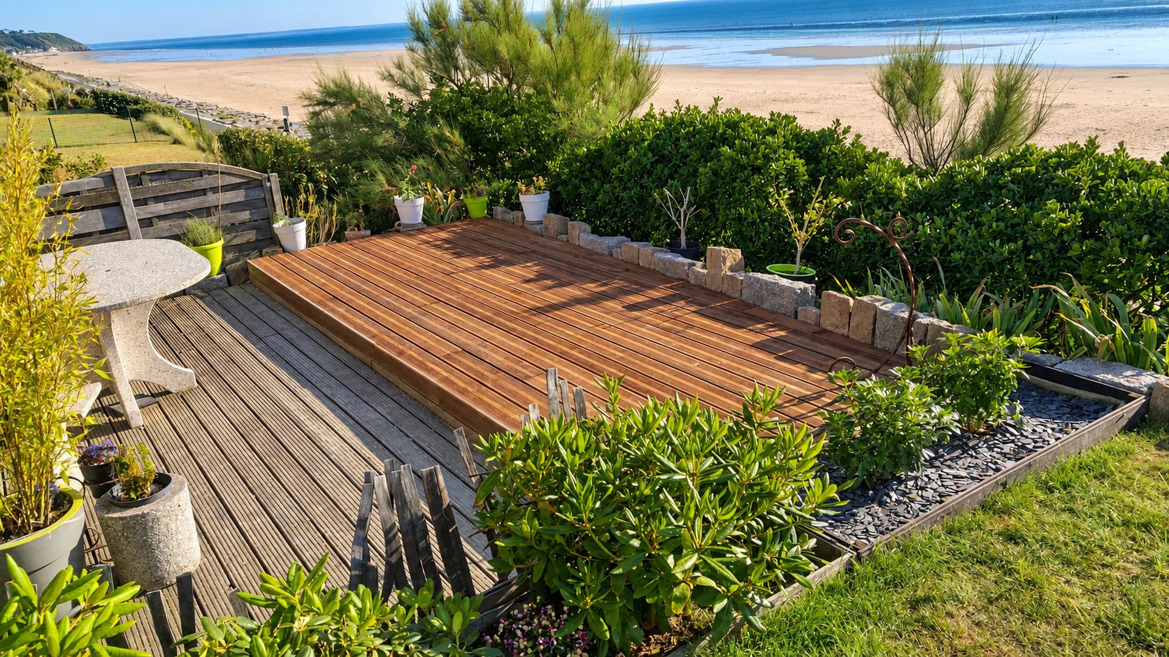 Une terrasse en bois à deux niveaux entourée de massifs, de pots de fleurs et d'un muret de pierres, avec la plage et la mer en arrière-plan.